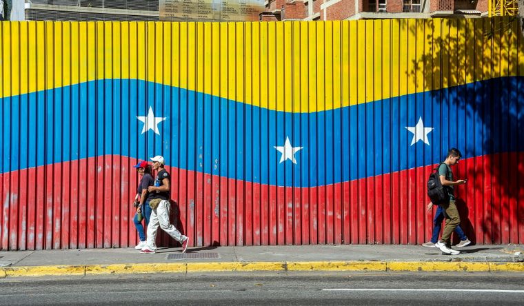Dos personas caminan delante de una bandera de Venezuela, país castigado por el poder de Estados Unidos. Foto Dpa&nbsp;