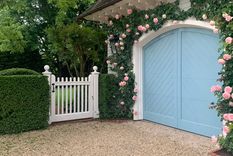 El azul pastel elegido para este garaje se complementa a la perfección con las rosas trepadoras de color rosa pastel que enmarcan las puertas arqueadas y la puerta blanca de estilo antiguo. Foto: Michael Derrig/Landscape Details