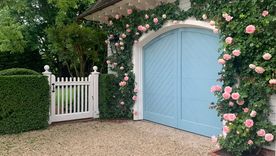 El azul pastel elegido para este garaje se complementa a la perfección con las rosas trepadoras de color rosa pastel que enmarcan las puertas arqueadas y la puerta blanca de estilo antiguo. Foto: Michael Derrig/Landscape Details