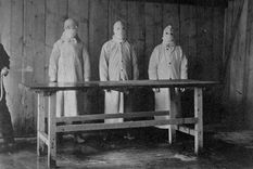 Preparaciones para una autopsia. Un asistente rocía ácido carbólico sobre una mesa antes de que tres médicos protegidos de pies a cabeza examinen un cuerpo.