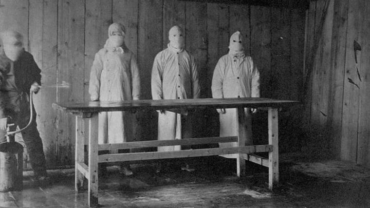 Preparaciones para una autopsia. Un asistente rocía ácido carbólico sobre una mesa antes de que tres médicos protegidos de pies a cabeza examinen un cuerpo.