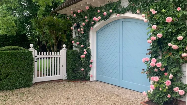 El azul pastel elegido para este garaje se complementa a la perfección con las rosas trepadoras de color rosa pastel que enmarcan las puertas arqueadas y la puerta blanca de estilo antiguo. Foto: Michael Derrig/Landscape Details